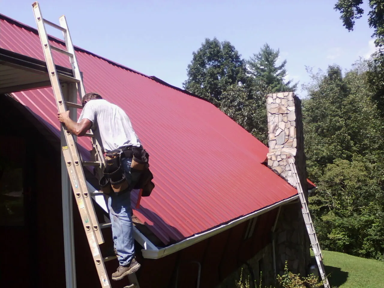 Roofer on a ladder beside a red metal roof and stone chimney — Soffit & Fascia Repair by Roofing Brothers in Land O Lakes